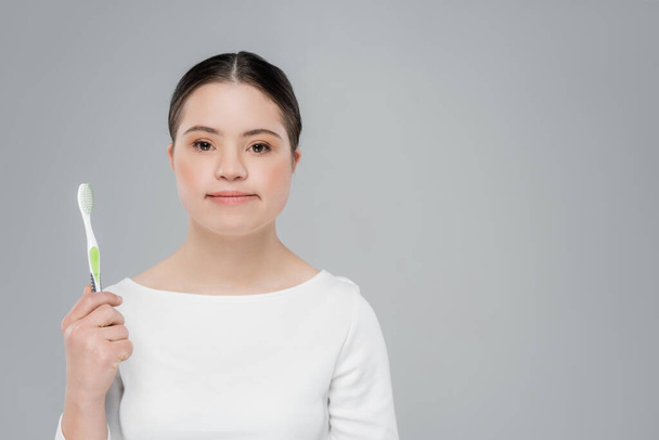 Young woman with down syndrome holding toothbrush and looking at camera isolated on grey  - Fotó, kép
