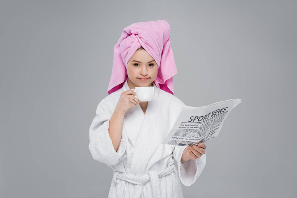 Woman with down syndrome in bathrobe holding cup and newspaper isolated on grey  - Photo, image
