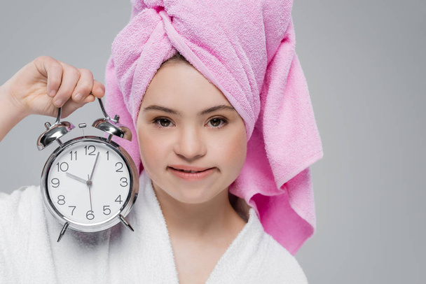 Woman with down syndrome in bathrobe and towel holding alarm clock isolated on grey  - Photo, Image