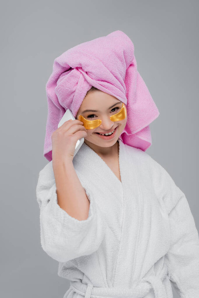 Smiling model with down syndrome in bathrobe and eye patches talking on smartphone isolated on grey  - Photo, Image