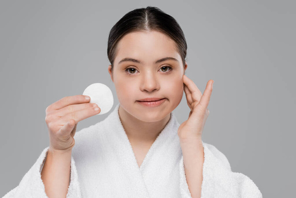 Woman with down syndrome in bathrobe holding cotton pad and touching face isolated on grey  - Fotó, kép