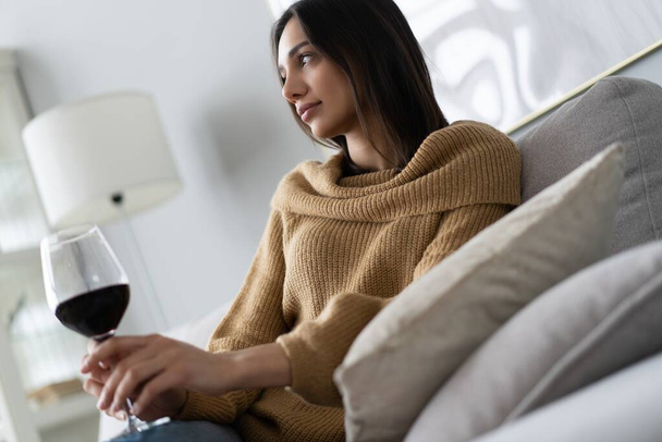 Красивая, привлекательная, стильная молодая женщина сидит на диване, поднимая бокал с красным вином, исследуя, пробуя напитки - Фото, изображение