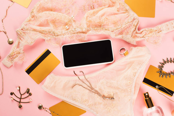 ランジェリー、クレジットカード、ピンクの背景にアクセサリーの近くに空白の画面を持つスマートフォンのトップビュー  - 写真・画像