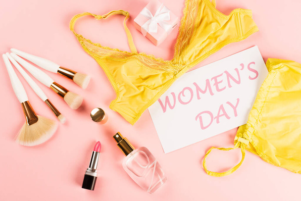 Top view of card with womens day lettering near bra, gift and cosmetic brushes on pink background  - Fotografie, Obrázek