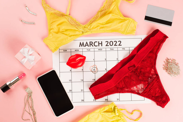 Вид сверху мартовского календаря у смартфона и нижнее белье на розовом фоне  - Фото, изображение