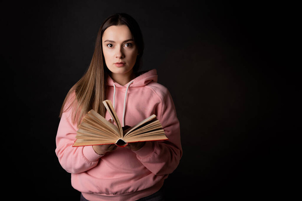 A young cute brunette in a pink hoodie, reading a book. Student with an open book in her hands, - Zdjęcie, obraz