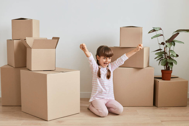 Indoor shot of happy positive little girl sitting on floor near carton boxes with personal piles during relocation to a new apartment, raised arms, celebrating moving. - Foto, Imagem