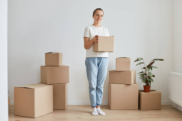 Full length portrait of attractive woman wearing white shirt and jeans posing at new apartment with carton boxes, looking at camera, holding box with her personal pile. - Valokuva, kuva