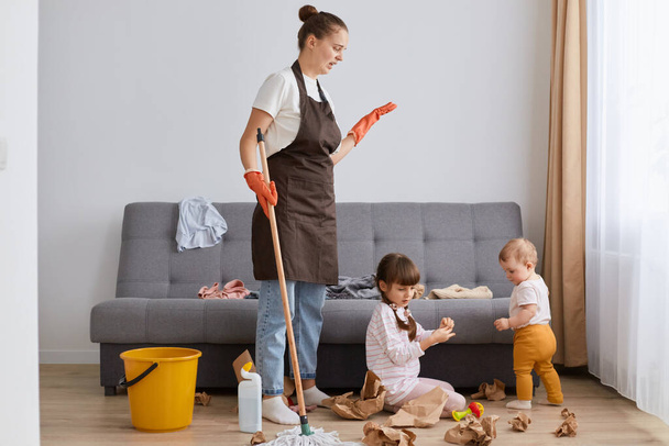 Shocked woman wearing white t shirt, brown apron and jeans, cleaning house, posing with her daughters, washing floor, upset that the kids have done a mess. - Photo, Image