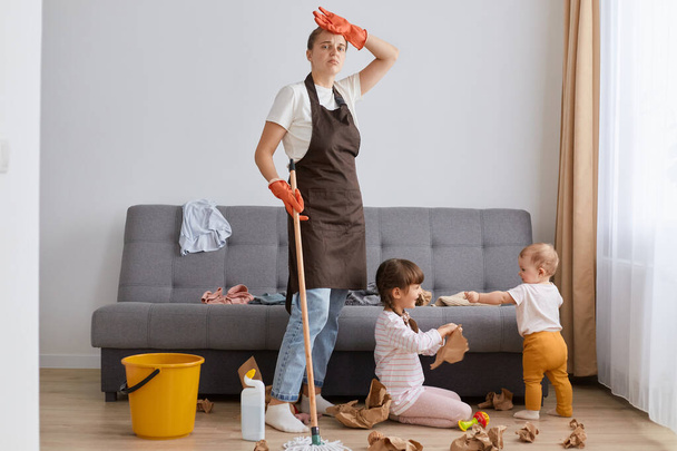 Tired woman wearing white t shirt, brown apron and jeans, cleaning house, posing with her children, keeping hand on her head, looks exhausted, needs to clean a mess her kids did. - Foto, afbeelding