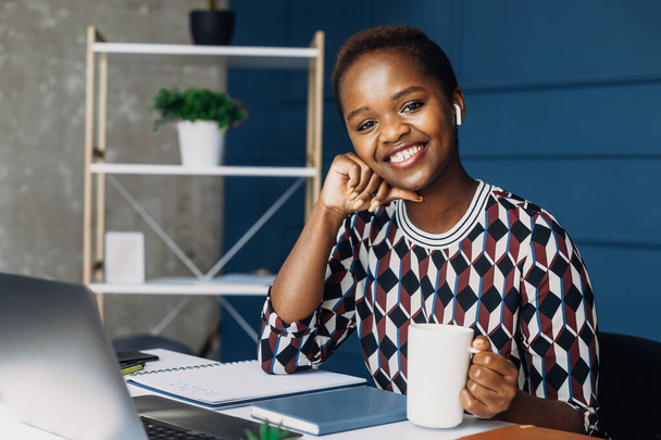 Ofis masasında poz veren ve elinde bir fincan kahve tutarken kameraya bakan Afrikalı bir kadının portresi. Şirket iş teknolojisi - Fotoğraf, Görsel