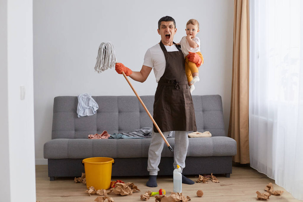 Angry aggressive man wearing casual attire and brown apron cleaning house with baby in hands, holding mop, looking at camera with shocked facial expression, being sad of mess. - Fotó, kép