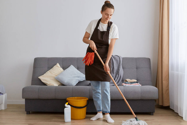 Full length portrait of dark haired woman wearing brown apron and casual clothing, washing floor with mop, doing household chores at home during weekend. - Foto, Imagen