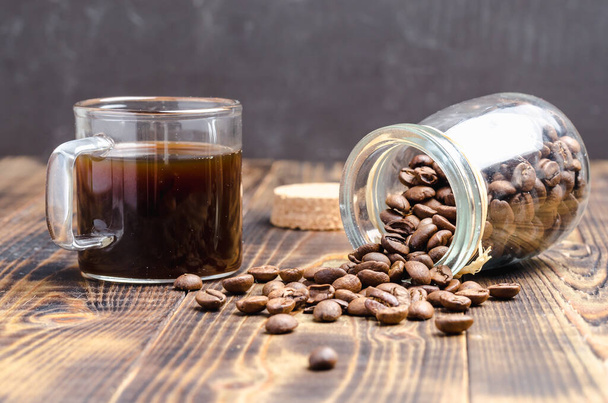 Cup of coffee and beans are scattered from a glass jar on a wooden table. - Valokuva, kuva