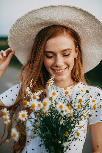 Portrait of a caucasian red-haired woman in hat holding a daisies bouquet. Closeup portrait. Beautiful young girl. Chamomile flower field. - Photo, Image