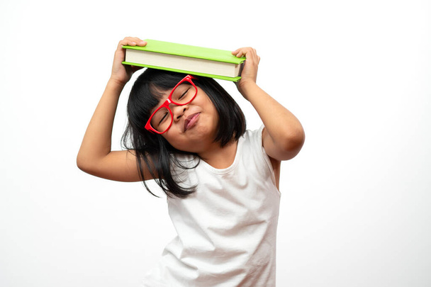 Дивна і щаслива азіатська маленька дошкільна дівчинка в червоних окулярах з зеленою книгою на голові, на білому ізольованому тлі. Концепція дитини в школі та освіта в початковій і дошкільній школі, домашній школі - Фото, зображення