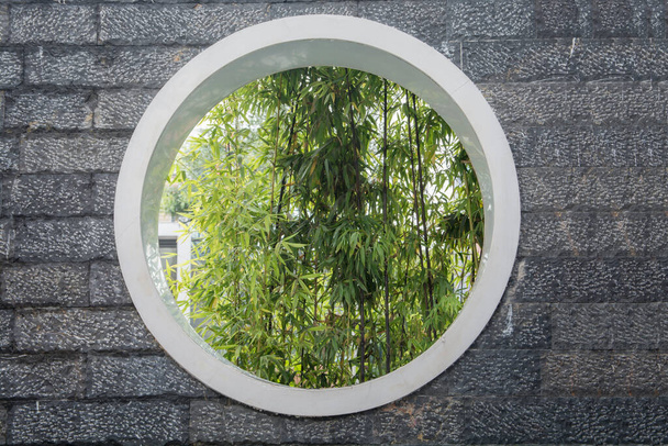 Bambusblätter durch ein großes Kreisfenster in einer Steinpflastermauer im Bezirk Pudong, Shanghai, China. - Foto, Bild