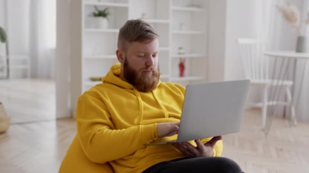 Bärtiger Mann mit Laptop arbeitet online und sitzt zu Hause - Filmmaterial, Video