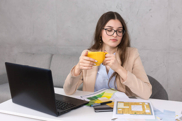 テーブルに座っている若いデザイナーの女の子がタブレットに描画されます。コンピュータ上でビジネス計算を行います。コーヒーのマグカップから飲む. - 写真・画像