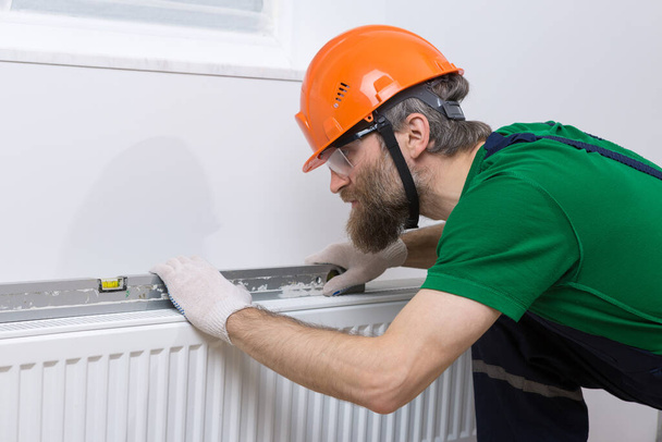 Um encanador masculino instala um radiador no sistema de aquecimento de um apartamento. Um tipo de macacão e uma chave de gás.. - Foto, Imagem