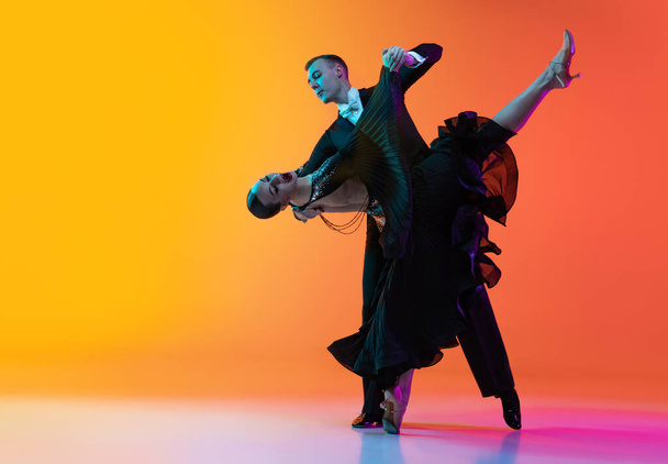 Full-length portrait of young beautiful man and woman dancing ballroom dance isolated over gradient orange pink background in neon light. Beauty, art, sport concept - Photo, Image
