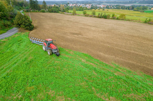A red tractor plows the land sown with green manure in the field, near the railway and the paved road. Agricultural industry. View from above. - Фото, зображення
