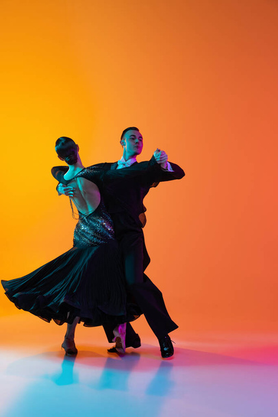 Full-length portrait of young beautiful man and woman dancing ballroom dance isolated over gradient orange pink background in neon light. Beauty, art, sport concept - Photo, Image