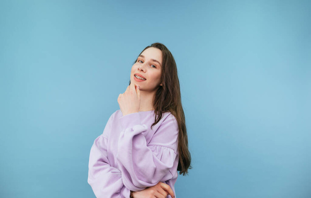 Happy girl in casual clothes posing at camera with a smile on her face on a blue background, looking at the camera and scratching her chin - Photo, Image