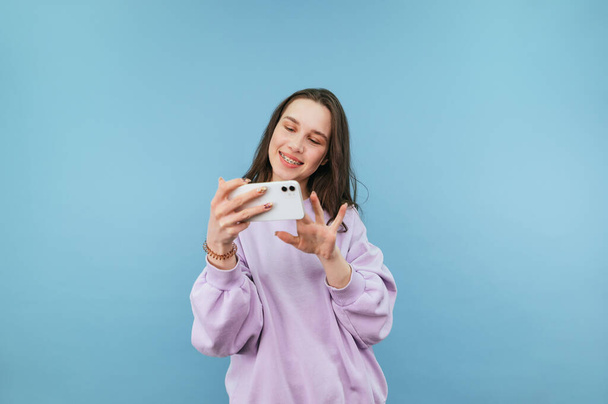 Приваблива дівчина в фіолетовому светрі стоїть зі смартфоном в руках на синьому фоні і грає в мобільні ігри на смартфоні з посмішкою на обличчі
. - Фото, зображення