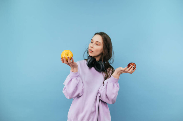 Привлекательная студентка в наушниках на шее стоит на синем фоне с яблоками в руках. - Фото, изображение