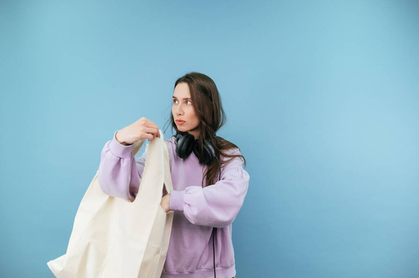Стильная девушка в наушниках на шее и повседневной одежде с эко-сумкой покупателя на плечах изолированы на синем фоне. - Фото, изображение
