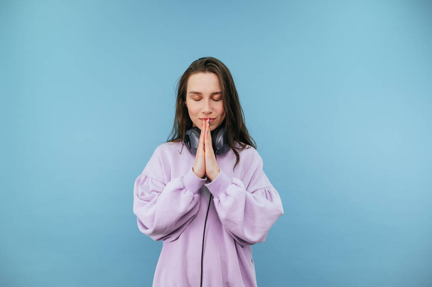 Beautiful student girl in headphones and casual clothes prays with closed eyes on a blue background. - Foto, Imagen