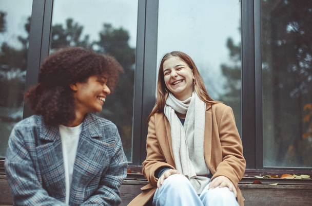 wo happy multiracial teen girls with takeway coffee sitting outdoors on fall day, talking and laughing. Улыбающиеся многонациональные лучшие подруги проводят время вместе на свежем воздухе осенью - Фото, изображение