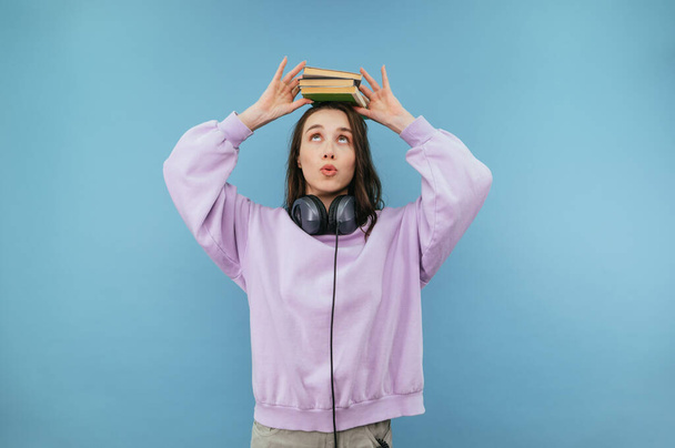 Csinos diák lány fülhallgató és rózsaszín pulóver tesz könyveket a fejére, és felnéz vicces arc, elszigetelt kék háttér - Fotó, kép
