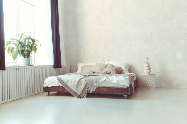 Stijlvolle minimalistische loft stijl slaapkamer interieur met lichte betonnen muren, kingsize houten bed met kussens, deken en metalen ronde nachtkastje met gedroogde bloemen in witte keramische vaas - Foto, afbeelding