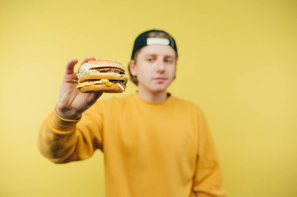 Young man in a bright sweatshirt shows the camera appetizing burger on a yellow background. Fast food concept - Photo, Image