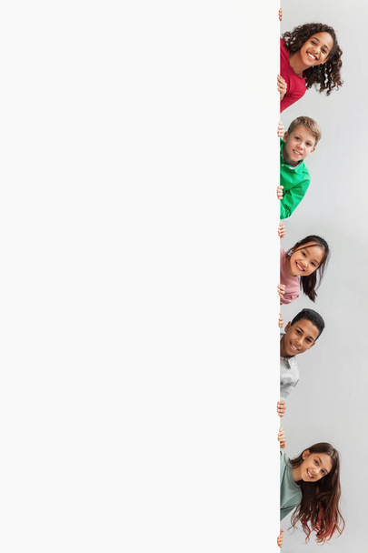 Niños multiétnicos escondidos detrás de la pizarra blanca sonriendo sobre fondo gris - Foto, Imagen