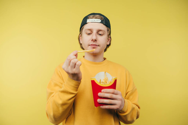 Cute guy in a cap and colored sweatshirt isolated on a yellow background with french fries in his hands. Young man having lunch of french fries. - Photo, Image
