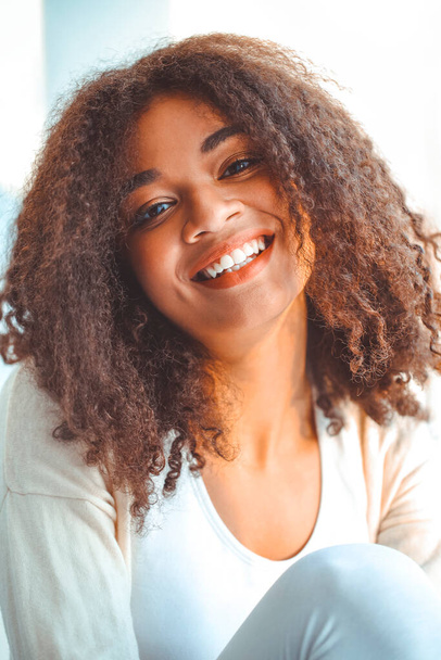 Pleased relaxed African American ethnicity girl in homewear sitting on windowsill against backdrop of curtains fluttering from light wind, happy afro female at home smiling gently with eyes closed - Φωτογραφία, εικόνα