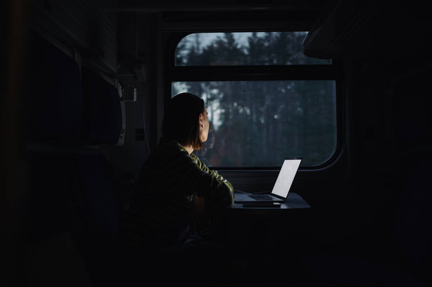 Žena sedí večer u stolu ve vlaku a pracuje na notebooku v kabelových sluchátkách, dívá se z okna na večerní výhledy. - Fotografie, Obrázek