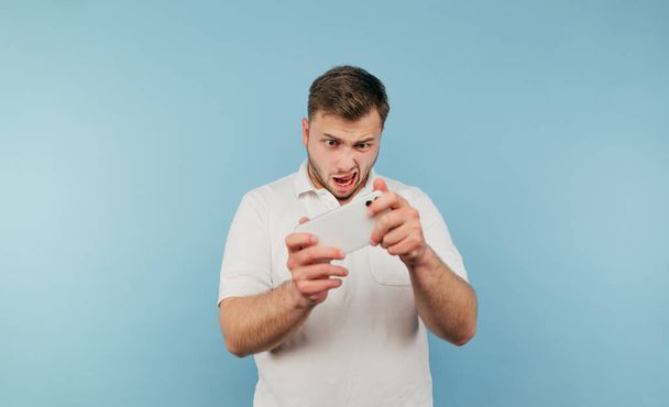 Αστείος συμπυκνωμένος άνθρωπος με τρίχες και λευκό T-shirt που παίζει παιχνίδια για κινητά στο smartphone σε μπλε φόντο. - Φωτογραφία, εικόνα