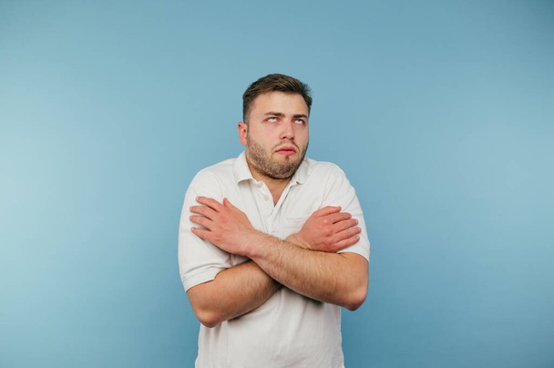 Замороженный мужчина в белой футболке, изолированной на синем фоне, обнимается и закатывает глаза. - Фото, изображение