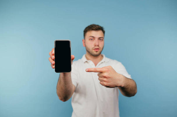 L'uomo serio con una maglietta bianca mostra un dito sullo schermo di uno smartphone in mano e guarda la fotocamera con un volto serio. Isolato. - Foto, immagini