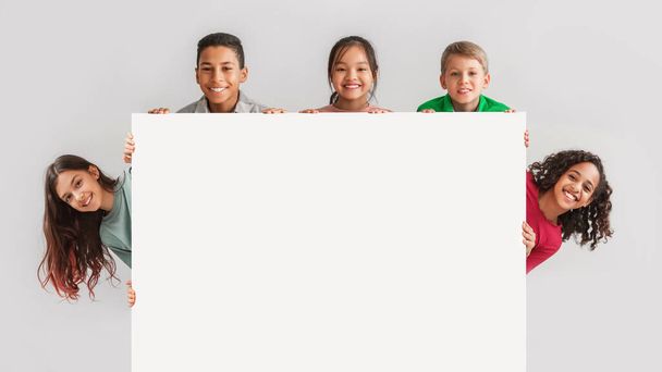 Onnellinen monipuolinen lapset piilossa takana tyhjä valkoinen lauta, harmaa tausta - Valokuva, kuva