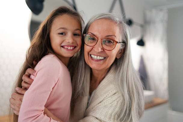 Senior grandmother and granddaughter standing indoors in bathroom, hugging and looking at camera. - Φωτογραφία, εικόνα
