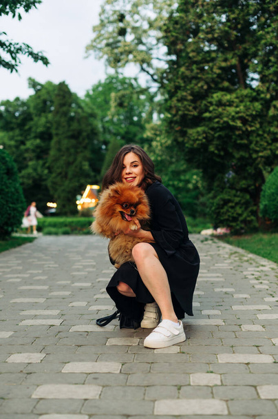 Ελκυστική χαμογελαστή γυναίκα με σακάκι κάθεται στο πάρκο με μια Pomeranian Spitz στα χέρια της και κοιτάζει την κάμερα και γελάει. - Φωτογραφία, εικόνα