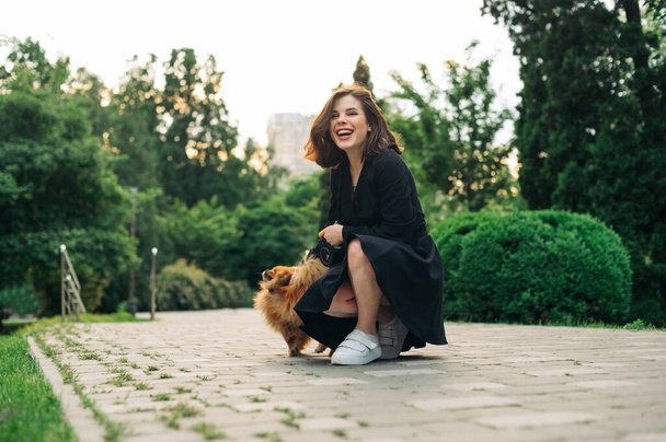 Positiivinen nainen koiran kanssa kävelyllä istuu jalkakäytävällä hymy kasvoillaan puiston taustalla.. - Valokuva, kuva