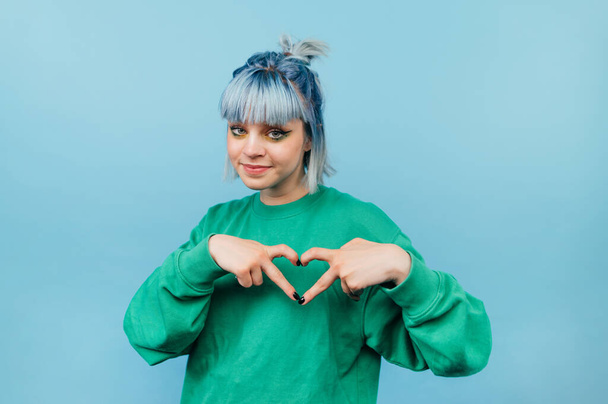 Belle adolescente dans un sweat-shirt vert et valse colorée montre un geste de coeur et regarde la caméra avec un sourire sur son visage. - Photo, image