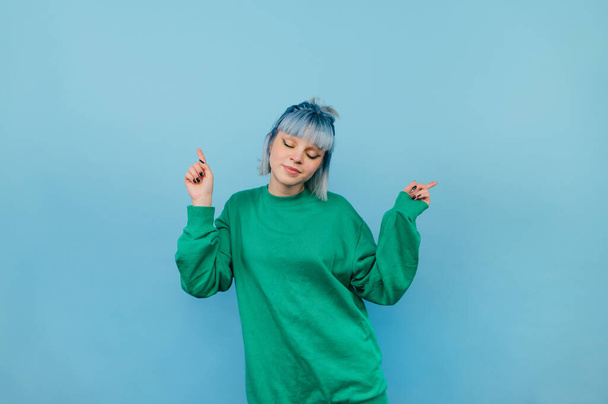 Cute bright girl in a green sweater dancing with closed eyes on a blue background with a smile on his face and having fun. - Photo, Image
