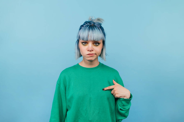 Chica hipster insatisfecha con el pelo de color señala un dedo en sí misma y mira infelizmente a la cámara sobre un fondo azul - Foto, imagen